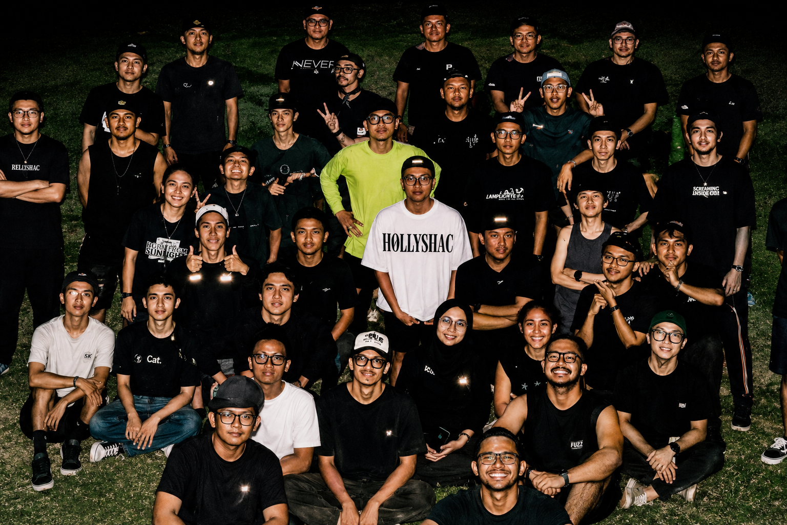 A large group of young men and women gathered on a grassy lawn at night, smiling for a group photo. In the center, a man stands out wearing a white oversized t-shirt with Hollyshac printed on the front.