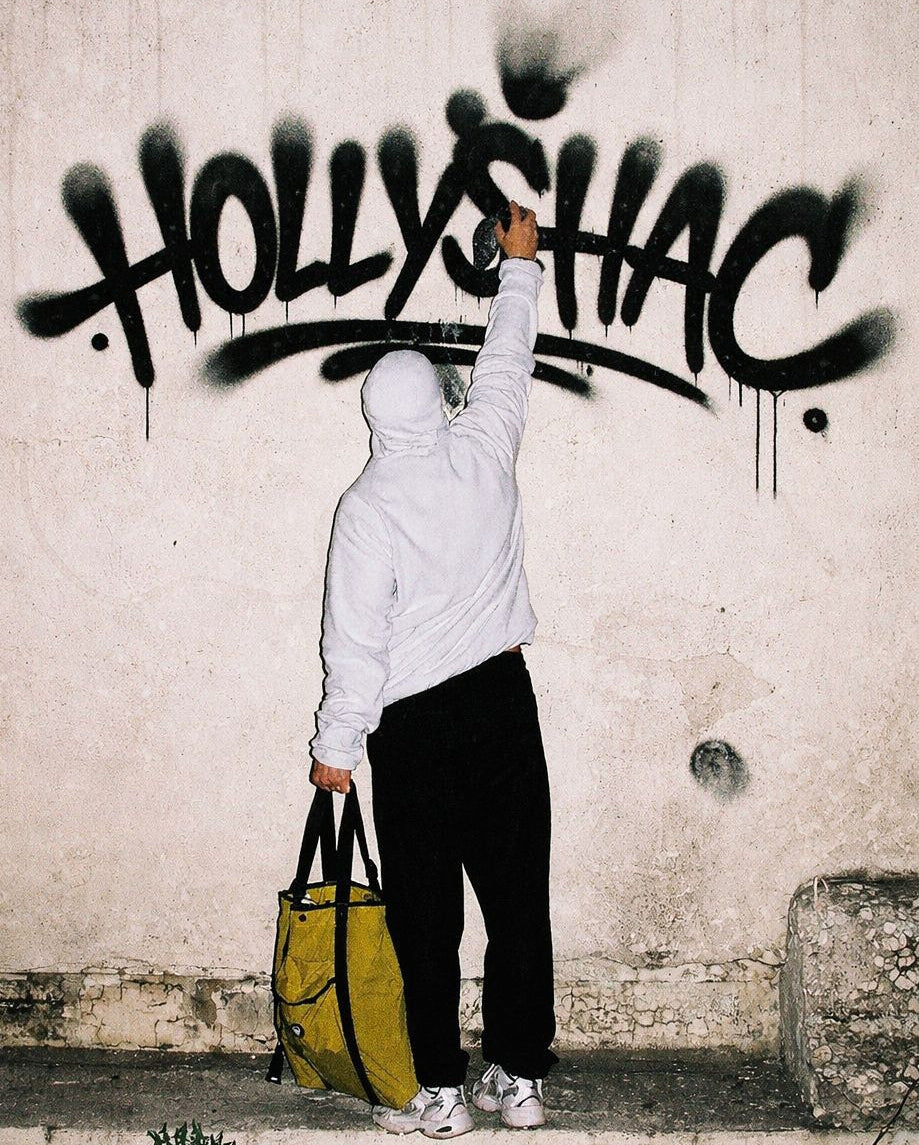 Street view of an artist in streetwear apparel using paint to tag the word Hollyshac across a concrete wall in an alleyway, showcasing authentic street lifestyle.